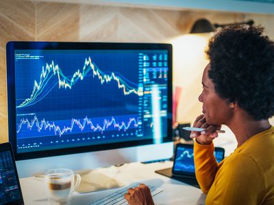 A woman sits at a desk with a cup of coffee in front of a large computer screen displaying financial stock charts. 