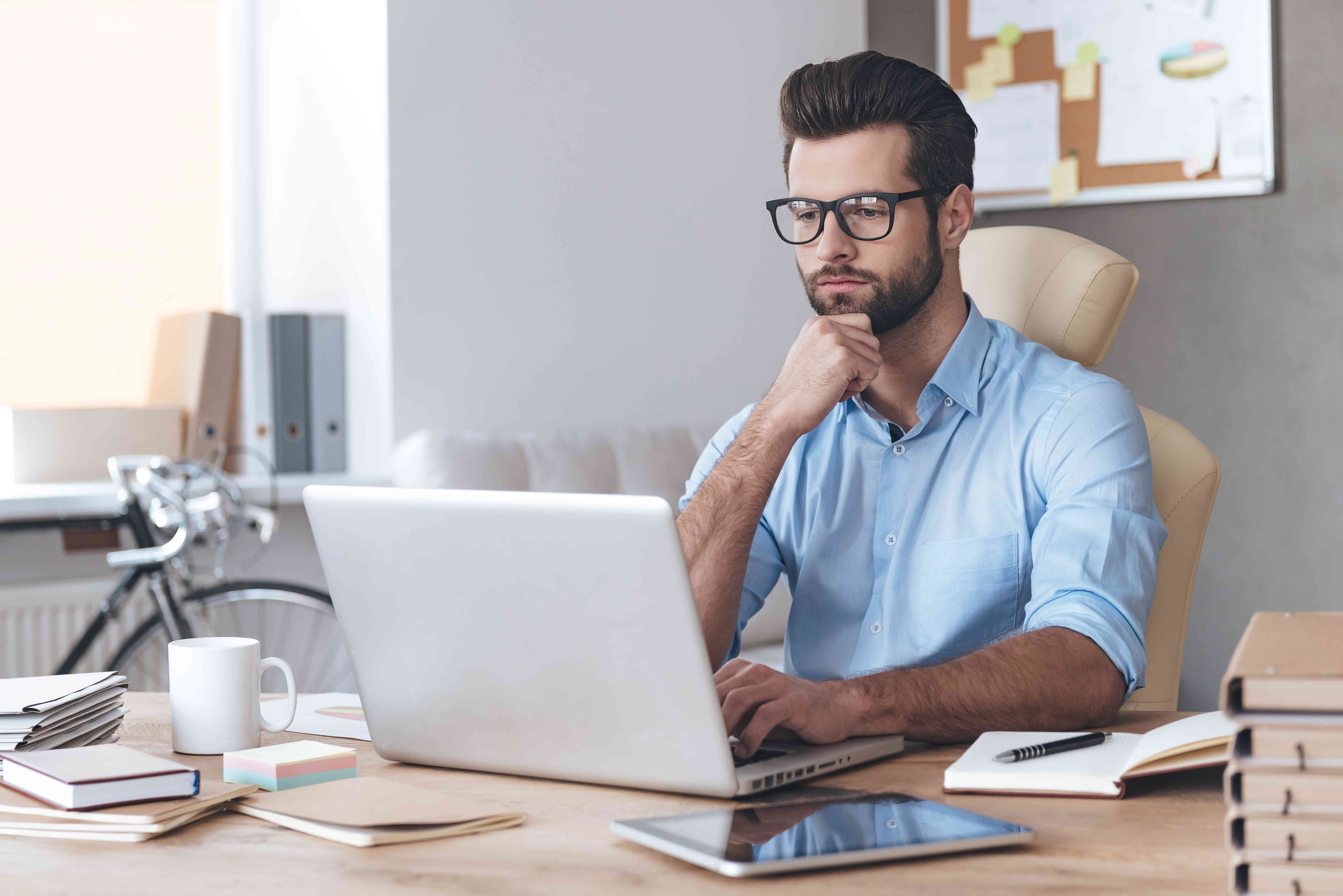 Young man sitting at home desk looking intently at his laptop screen