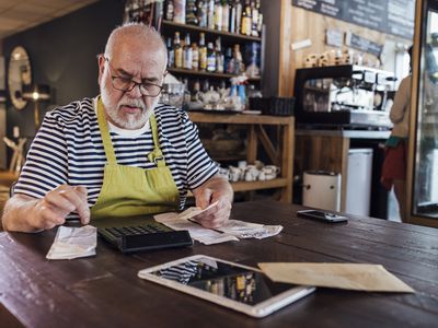 A medium shot of a man sitting at a table in his cafe adding up receipts on a calculator