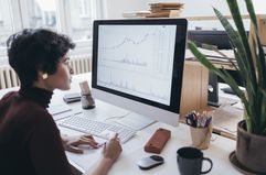 A woman sits at a desk, looking at a computer screen displaying stock figures and charts.