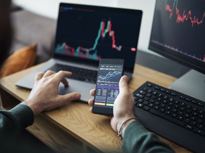 A man holds a cell phone displaying financial charts while sitting in front of two computers at a desk.