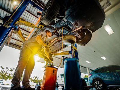 An auto mechanic performs a repair underneath an automobile hoisted on a garage lift.