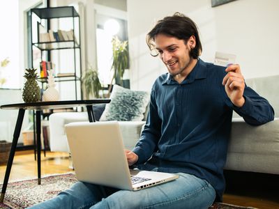 A man sits on the floor with his laptop, smiling and holding a payment card.