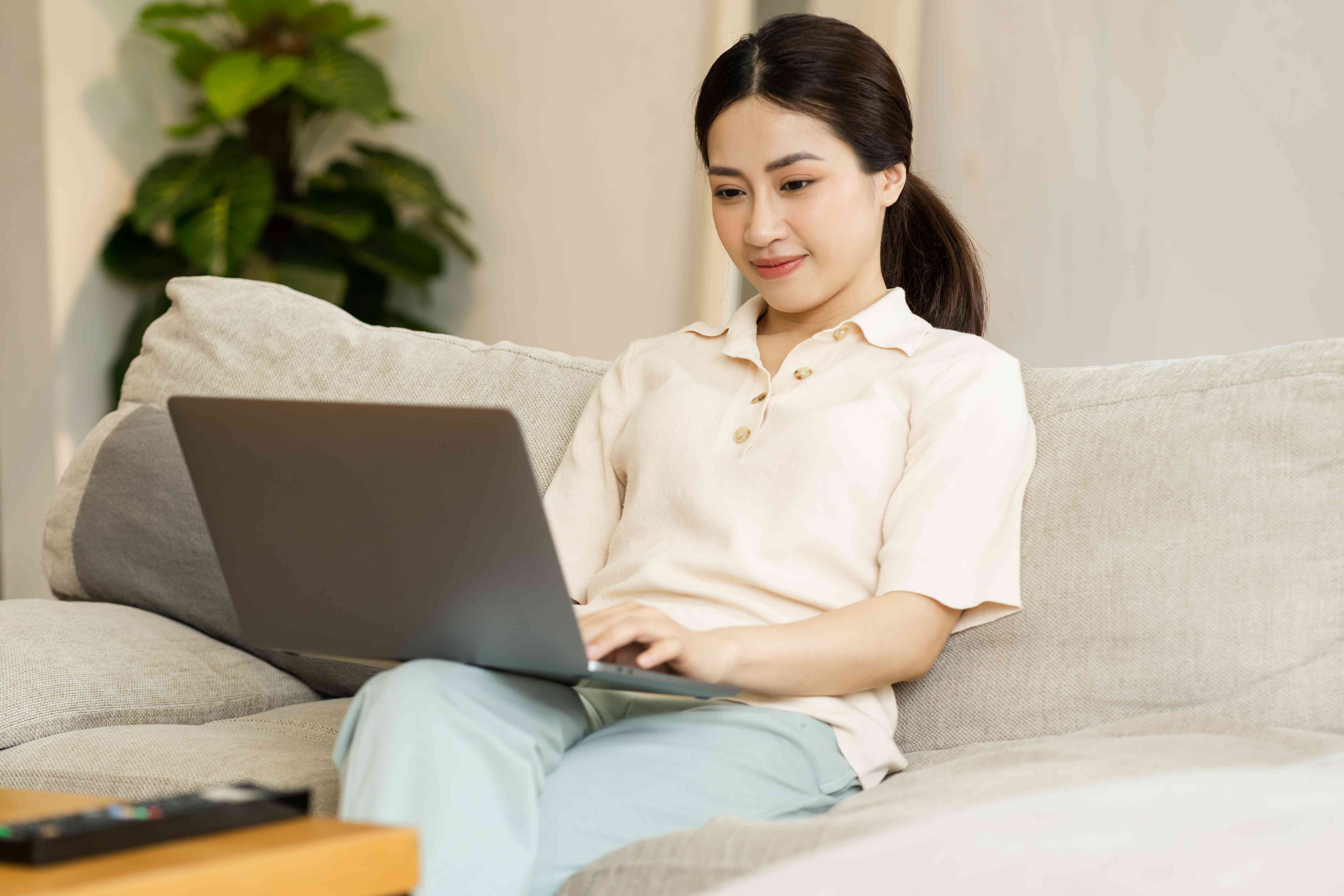 Young woman sitting on a her living room couch and smiling as she looks at something on her laptop