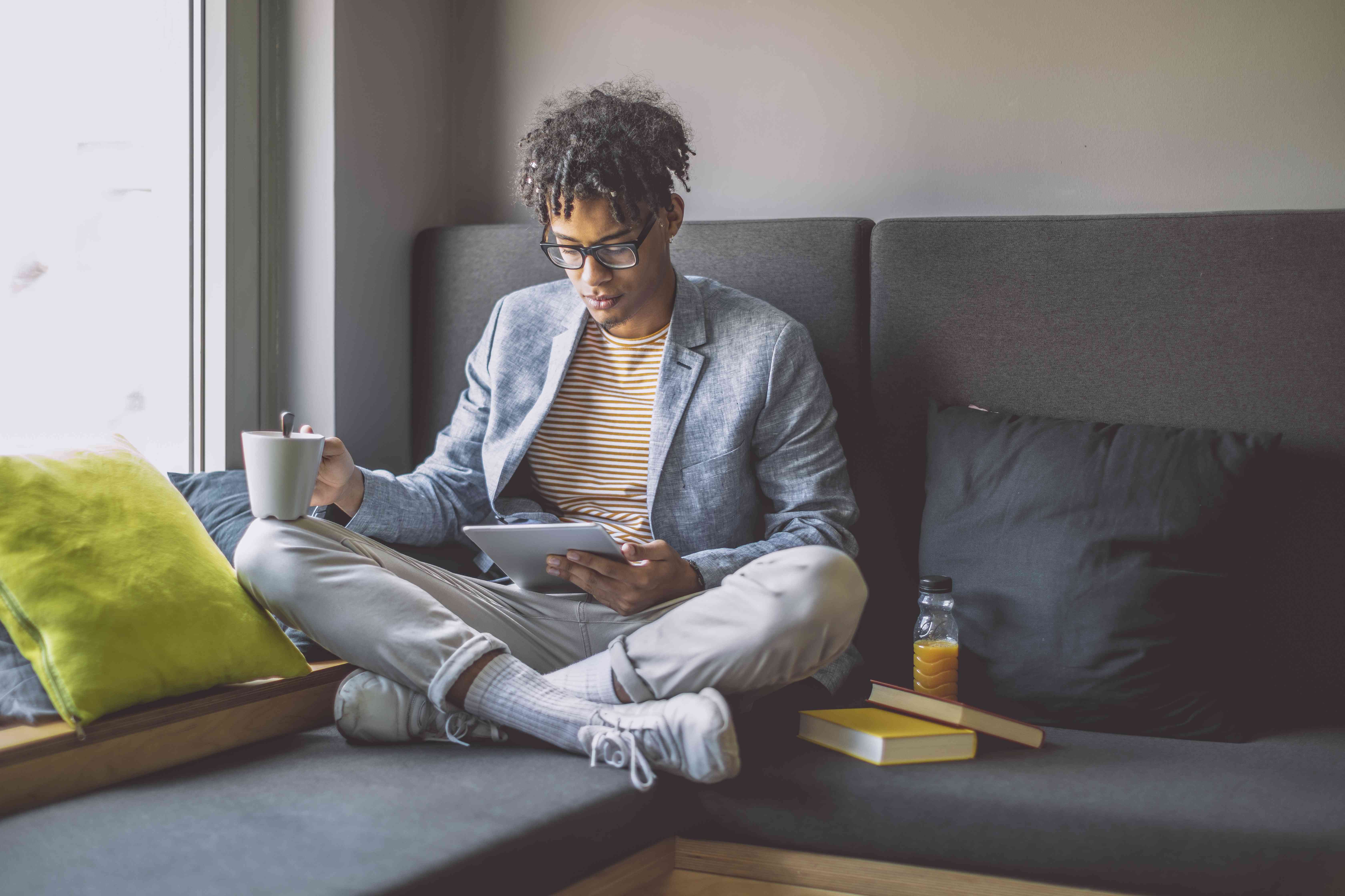 Young man sitting at home on his couch, looking intently at a table with a coffee cup in his other hand.