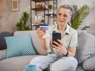 A woman sits on a couch, holding a bank card and mobile phone.