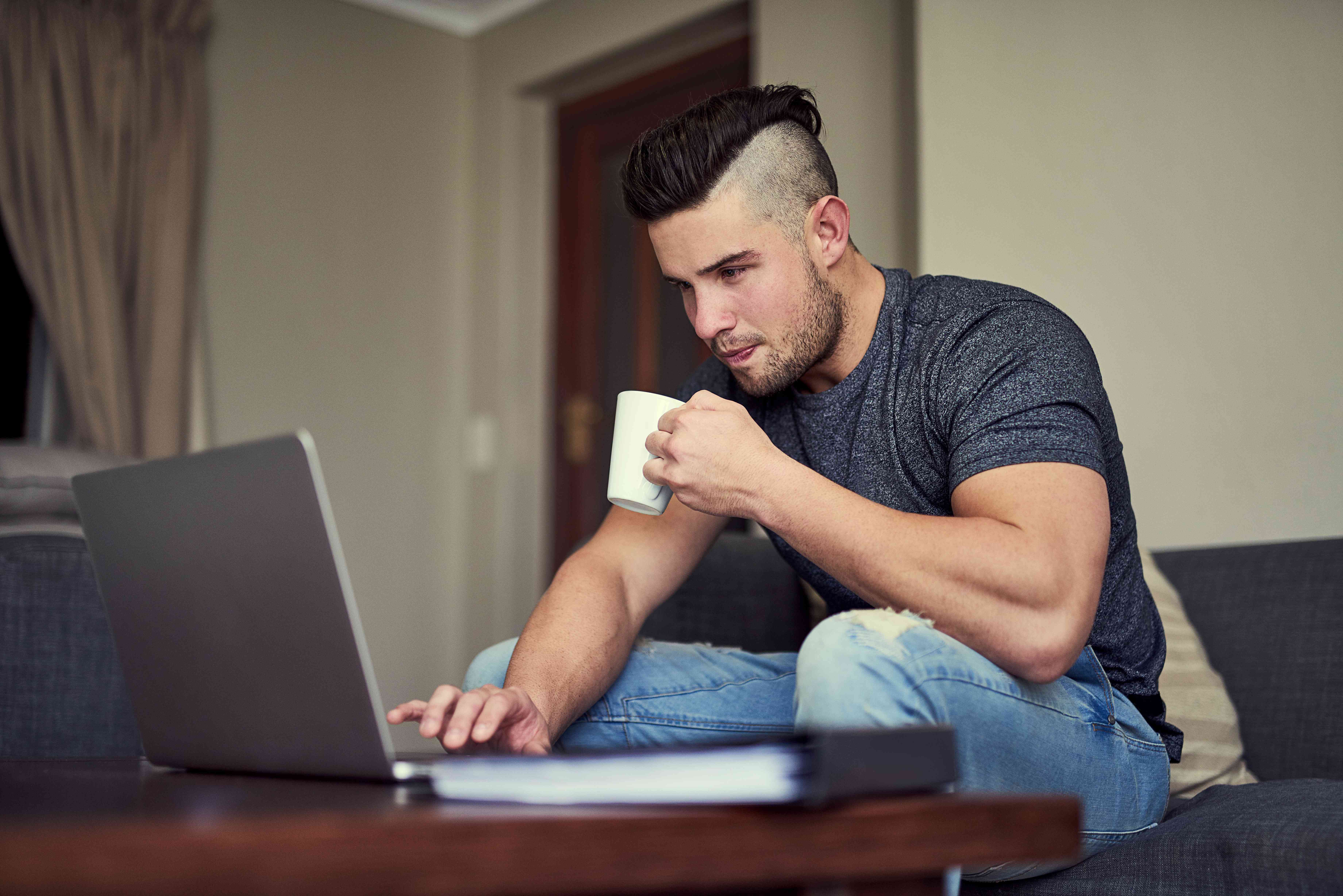 A man sits on a couch holding a mug while looking at a laptop that sits on the coffee table