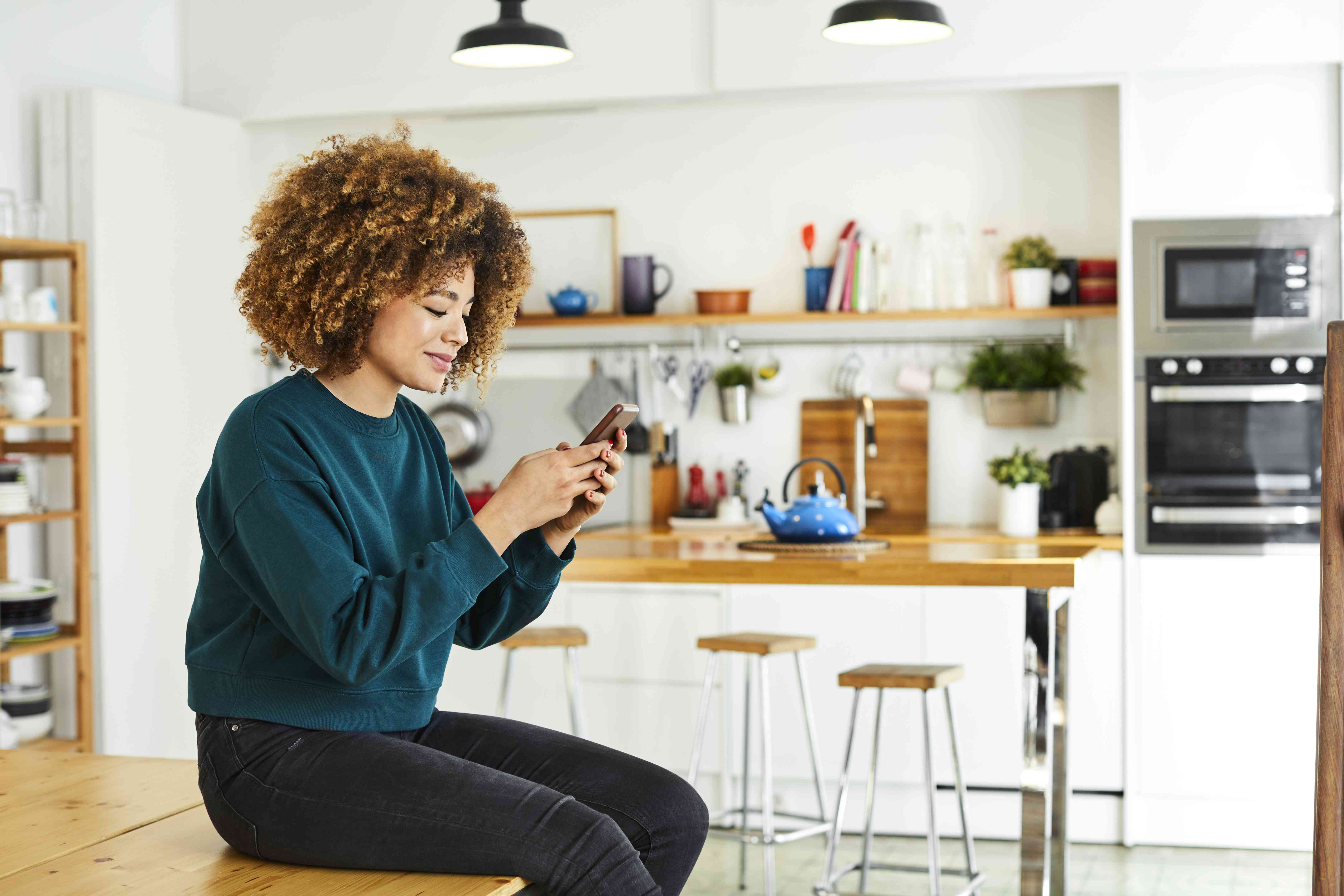 A woman in a teal sweater sits on a table in a kitchen looking at her smartphone