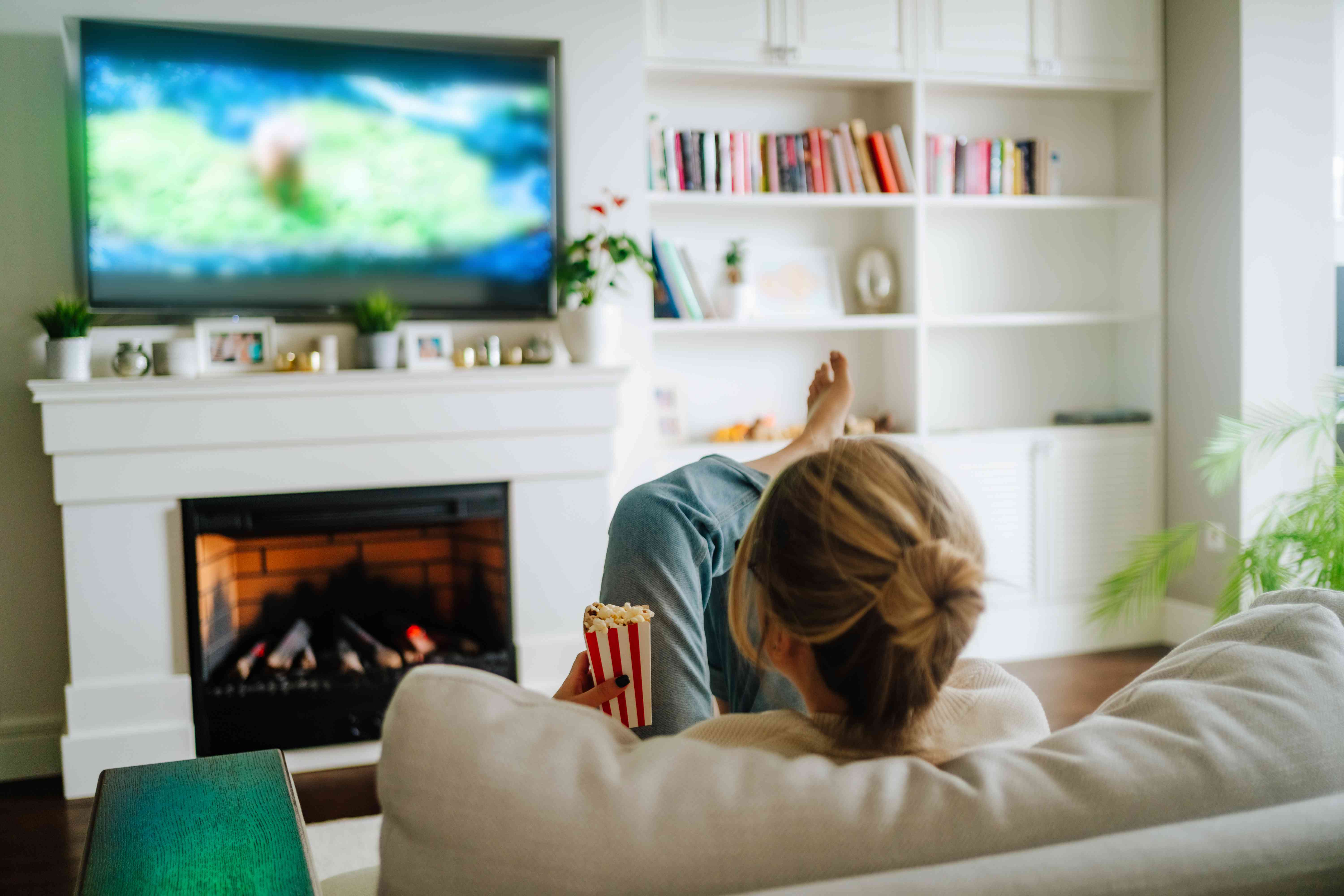 A woman watches TV on a couch while eating popcorn near a fireplace. 