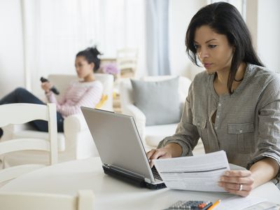 A woman seated at a kitchen table holds a document and peers at a laptop screen. In the background a young woman relaxes in an easy, television remote control in hand.