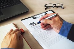 cropped hand of individual going over a resume with pen in hand, glasses on desk and a laptop