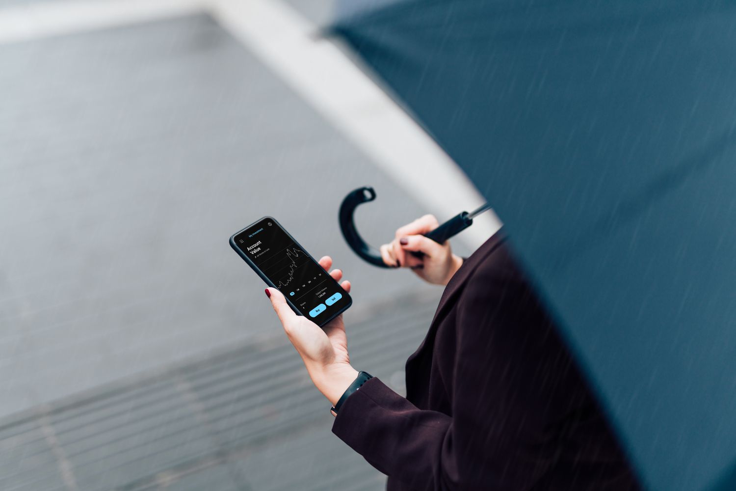 An investor standing outside and holding an umbrella while looking at weather futures investment data on their smartphone.