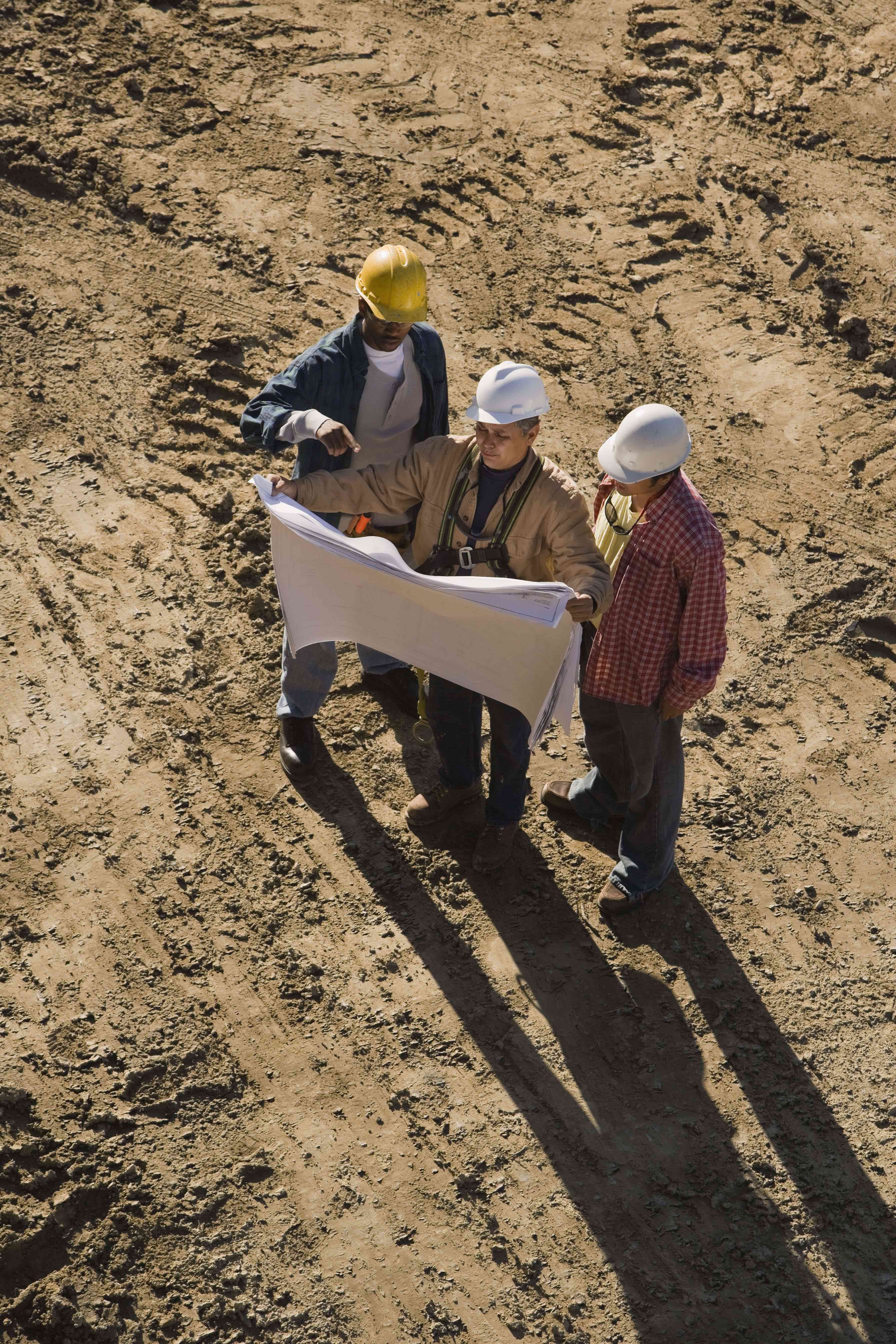 An engineer, an architect, and an earthmoving equipment operator look at a set of site plans while standing in the center of a construction site.