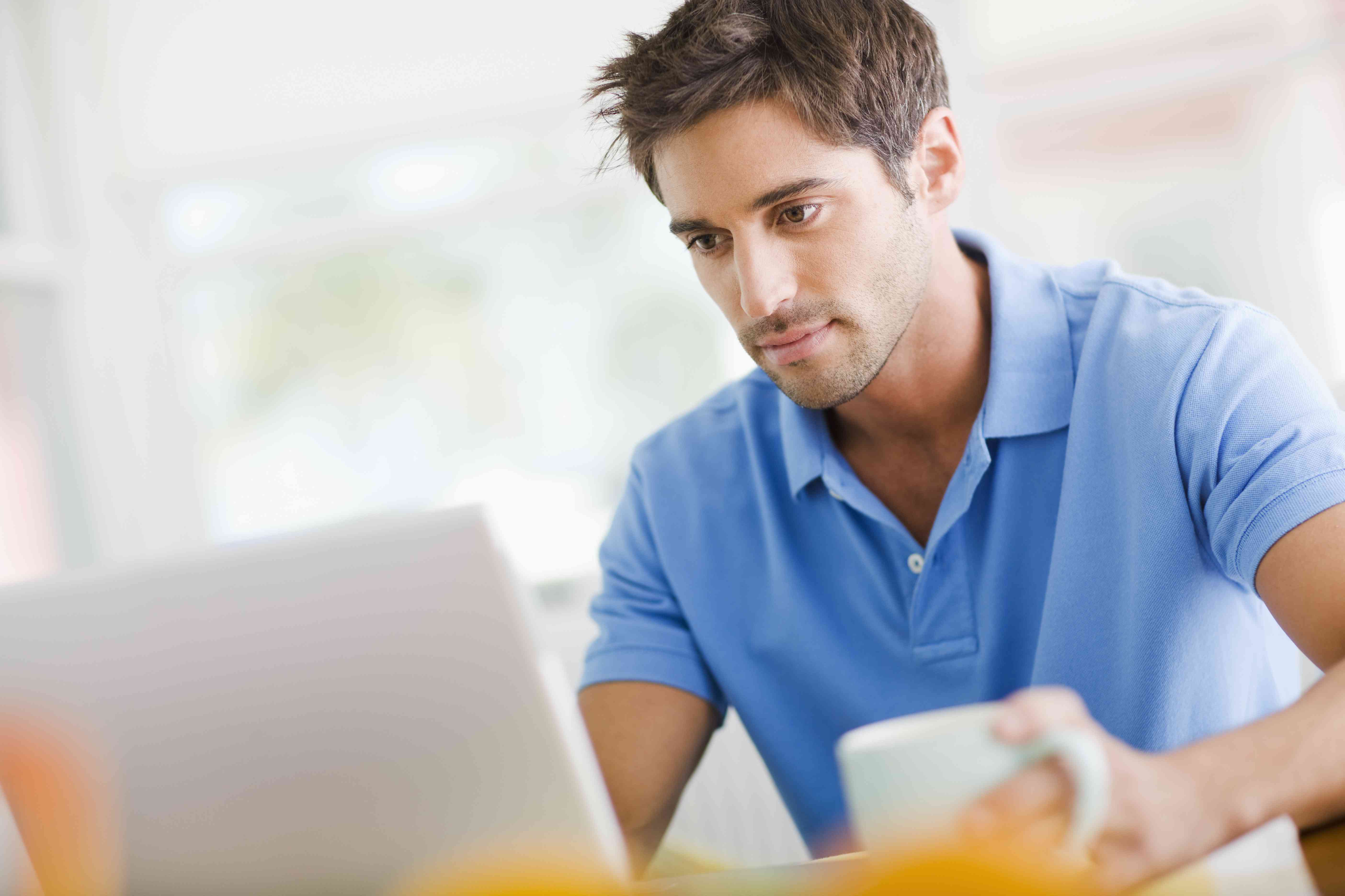 Young man at home, holding a coffee cup and looking at something on his laptop