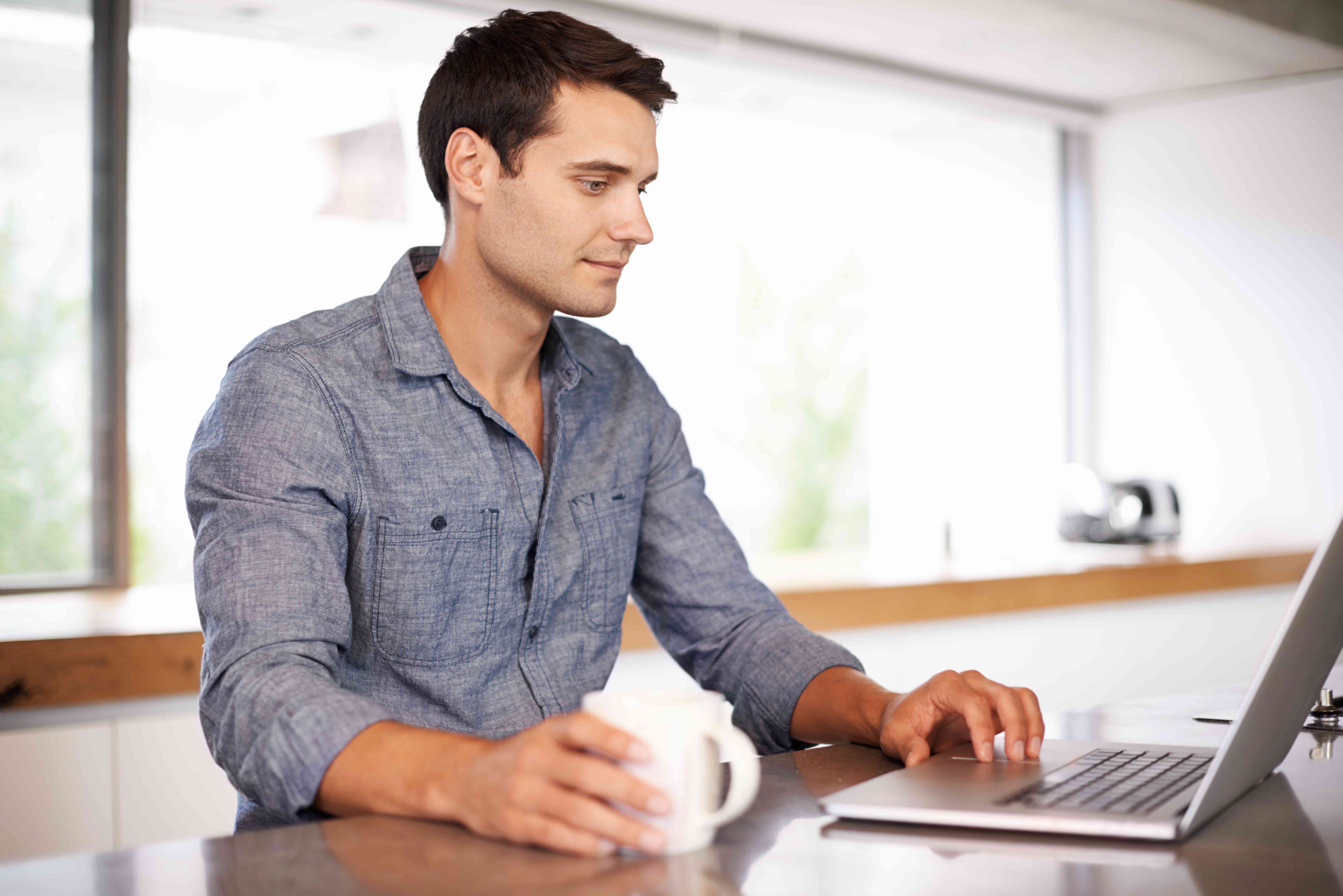 Young man at his kitchen counter with a coffee, looking at his laptop with a slight smile