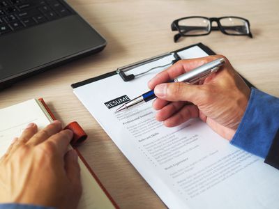 cropped hand of individual going over a resume with pen in hand, glasses on desk and a laptop