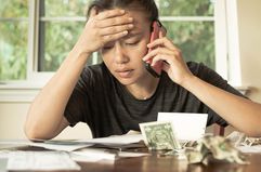 A stressed woman looks at bills on a table, holding a cell phone to her ear.