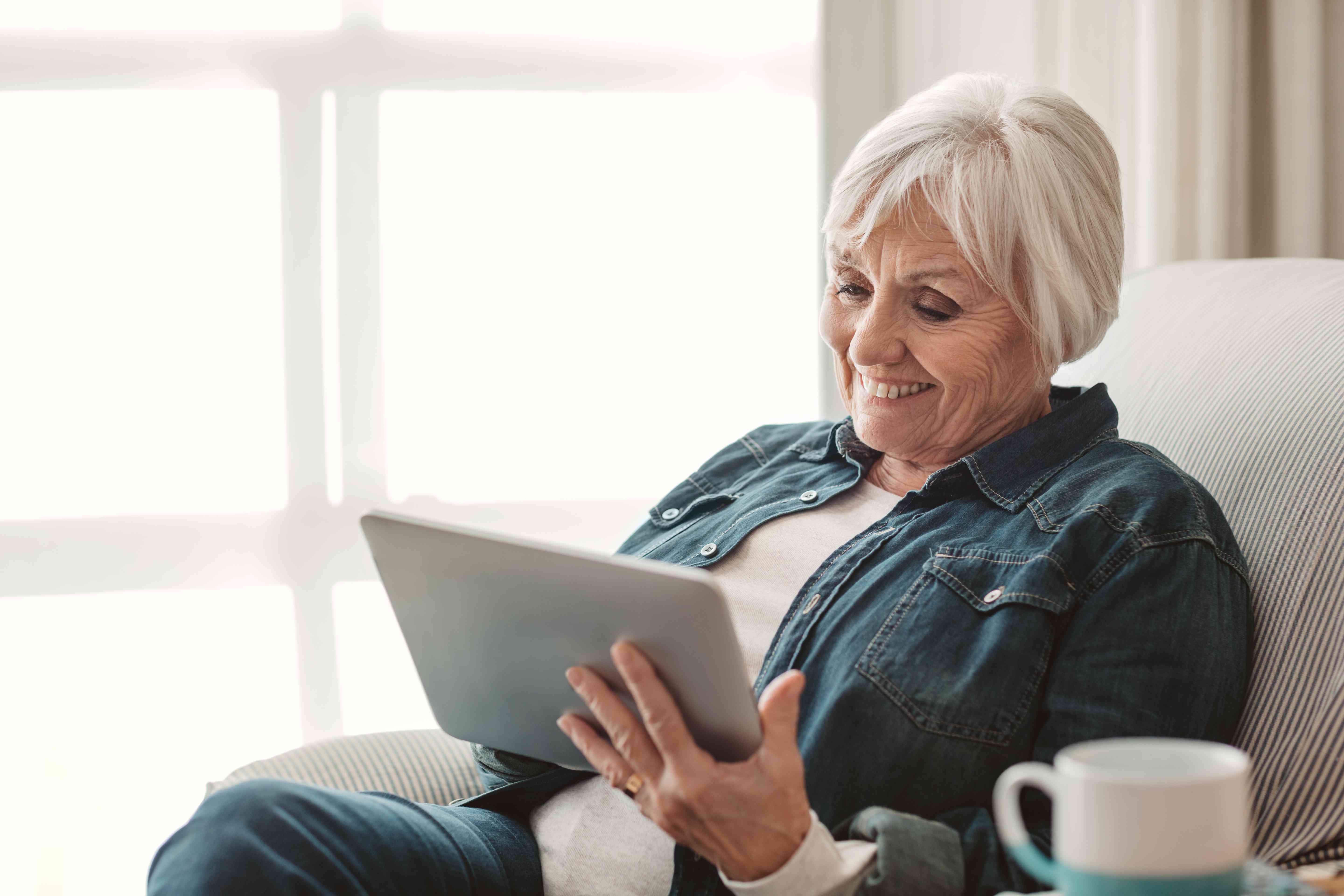 Older woman smiling as she relaxes on a chair in her living room while looking at a tablet