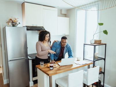 A couple standing together in a well-lit kitchen discuss something on a laptop screen.