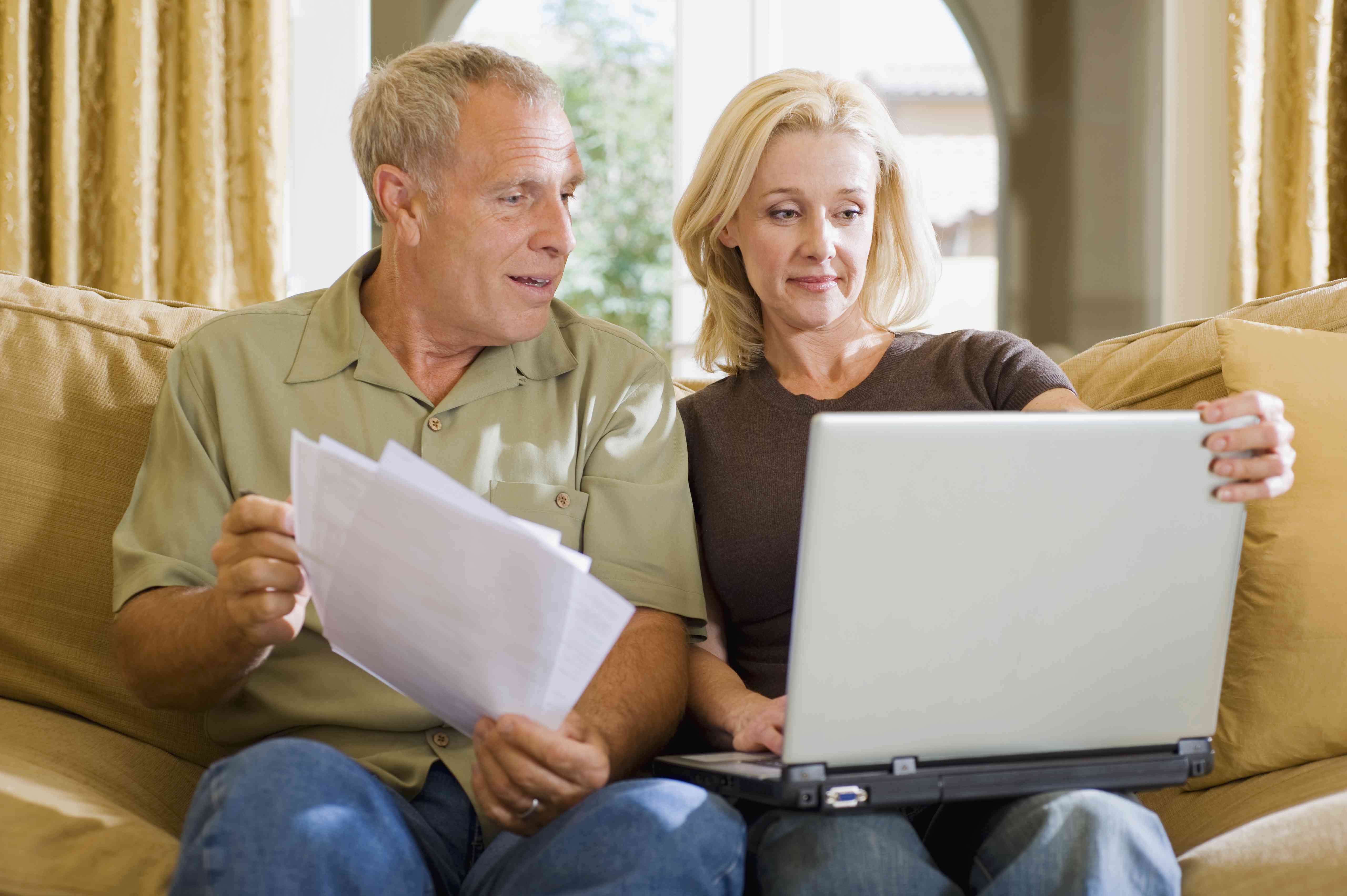 Older couple on living room couch, looking together at a laptop and financial documents