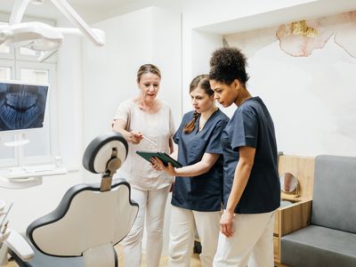 In a dental exam room, a woman reviews information on a tablet computer with a dentist and an assistant.