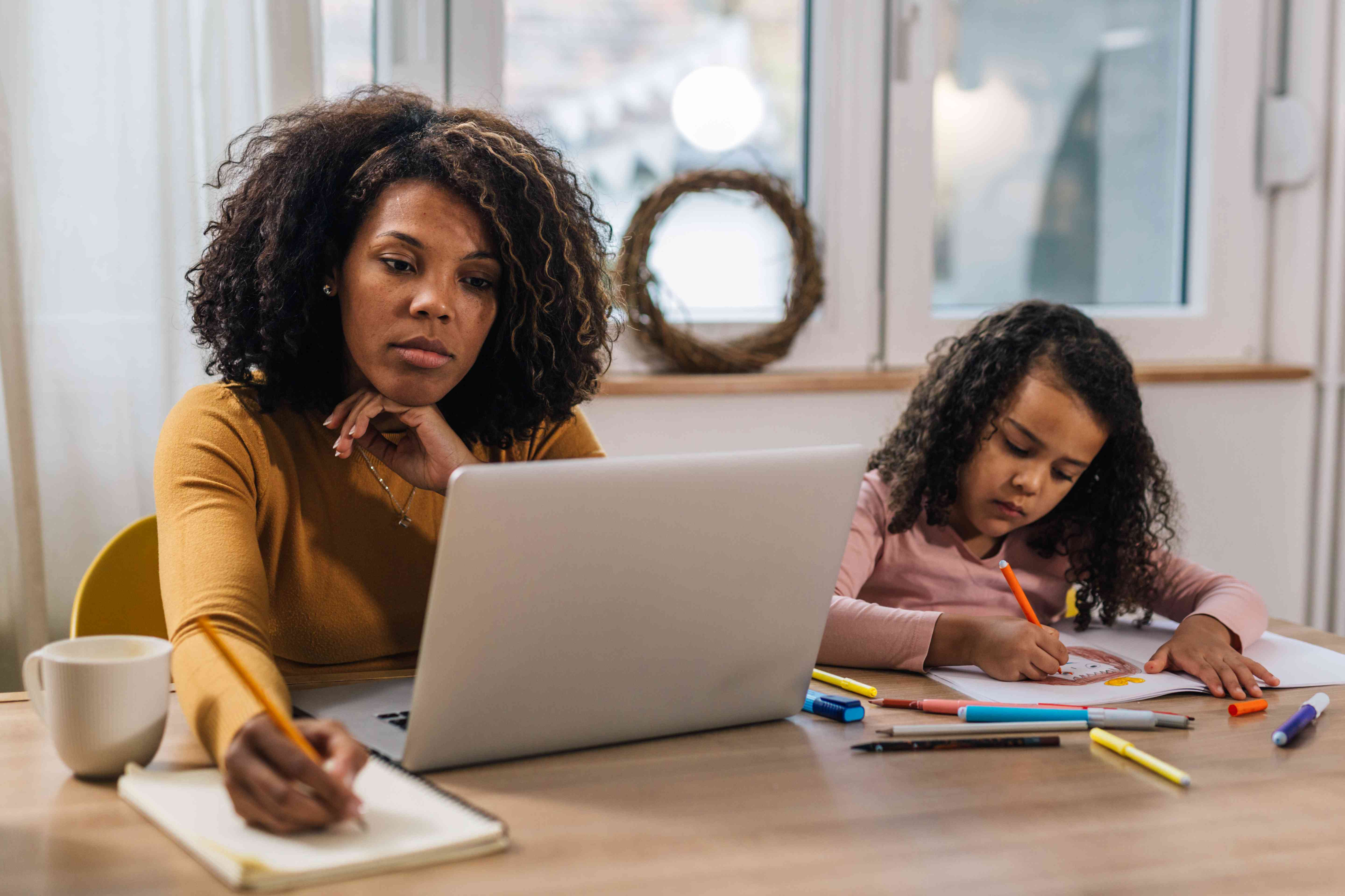 Mom at the kitchen table, studying something on her laptop while her daughter colors next to her