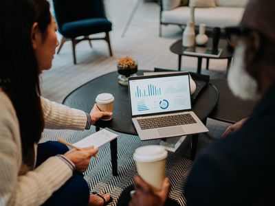 Two people sit on a sofa. They share a laptop, both looking at the statistics displayed, while they drink coffee.