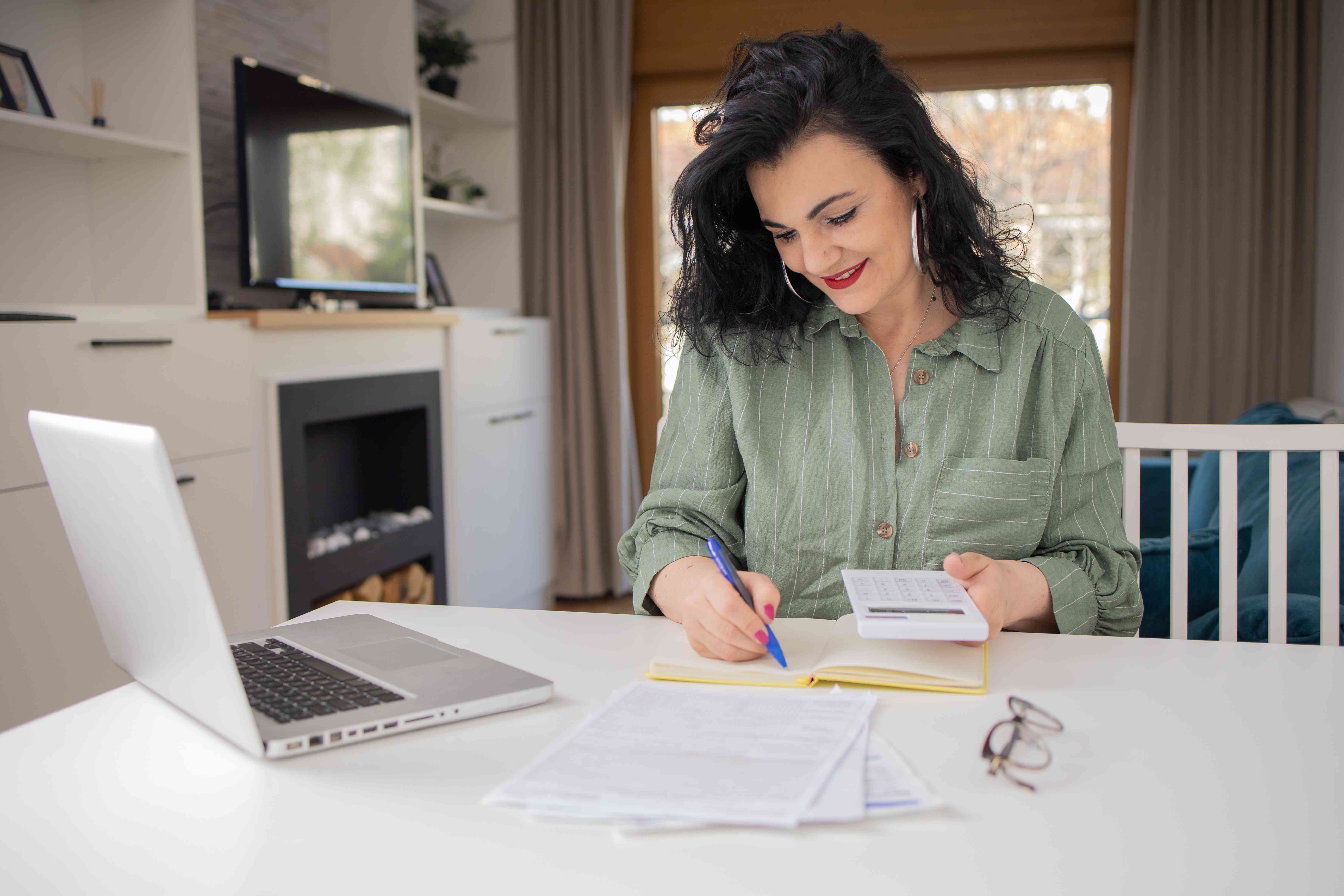 A person sitting in the living room with calculator in one hand and noting down details on notebook about the best options for free checking account with the other hand.