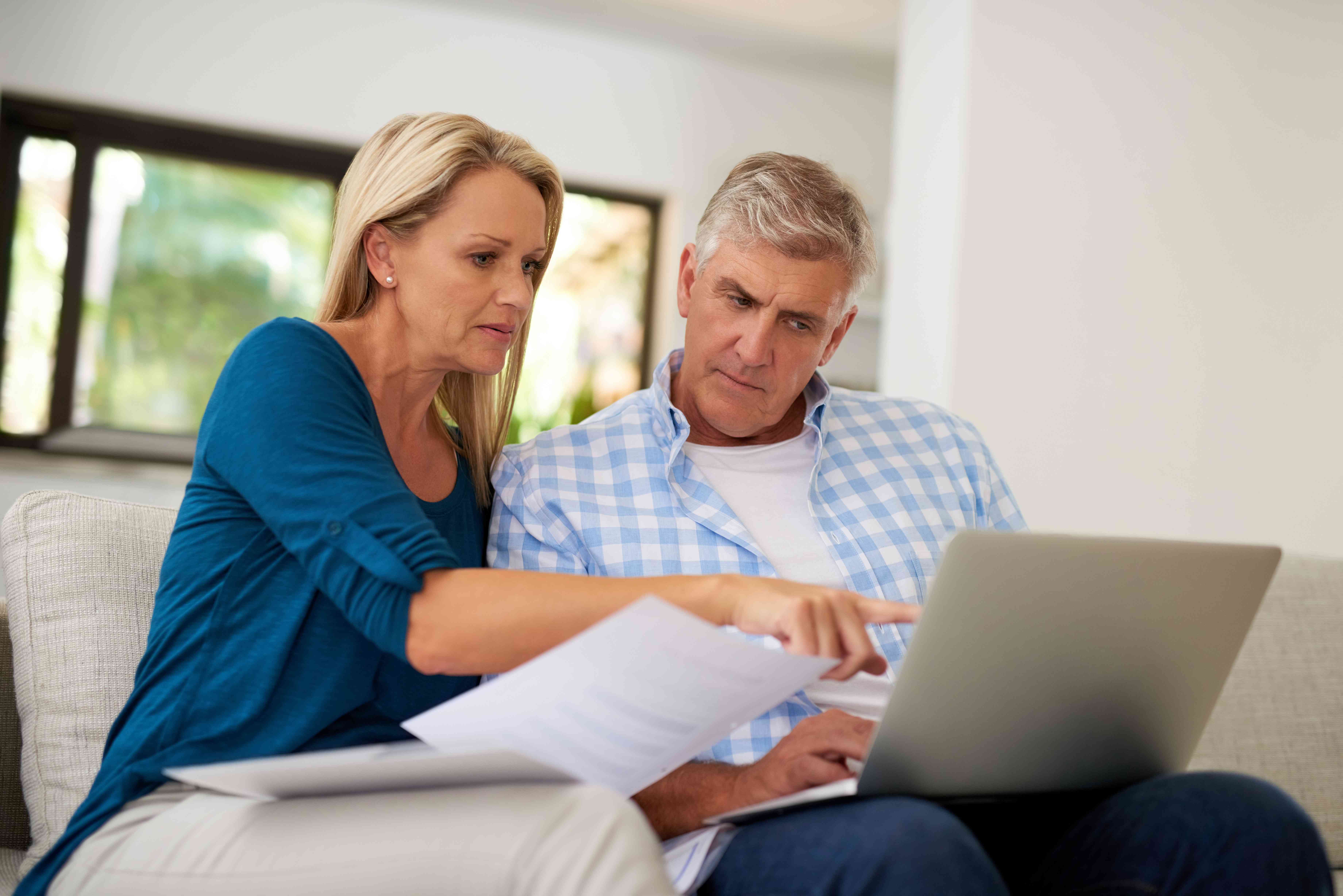 Older couple at home on living room couch looks intently at laptop and documents