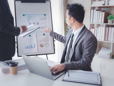 An accountant points to a display board holding charts and explains quarterly company earnings after tax during a meeting.