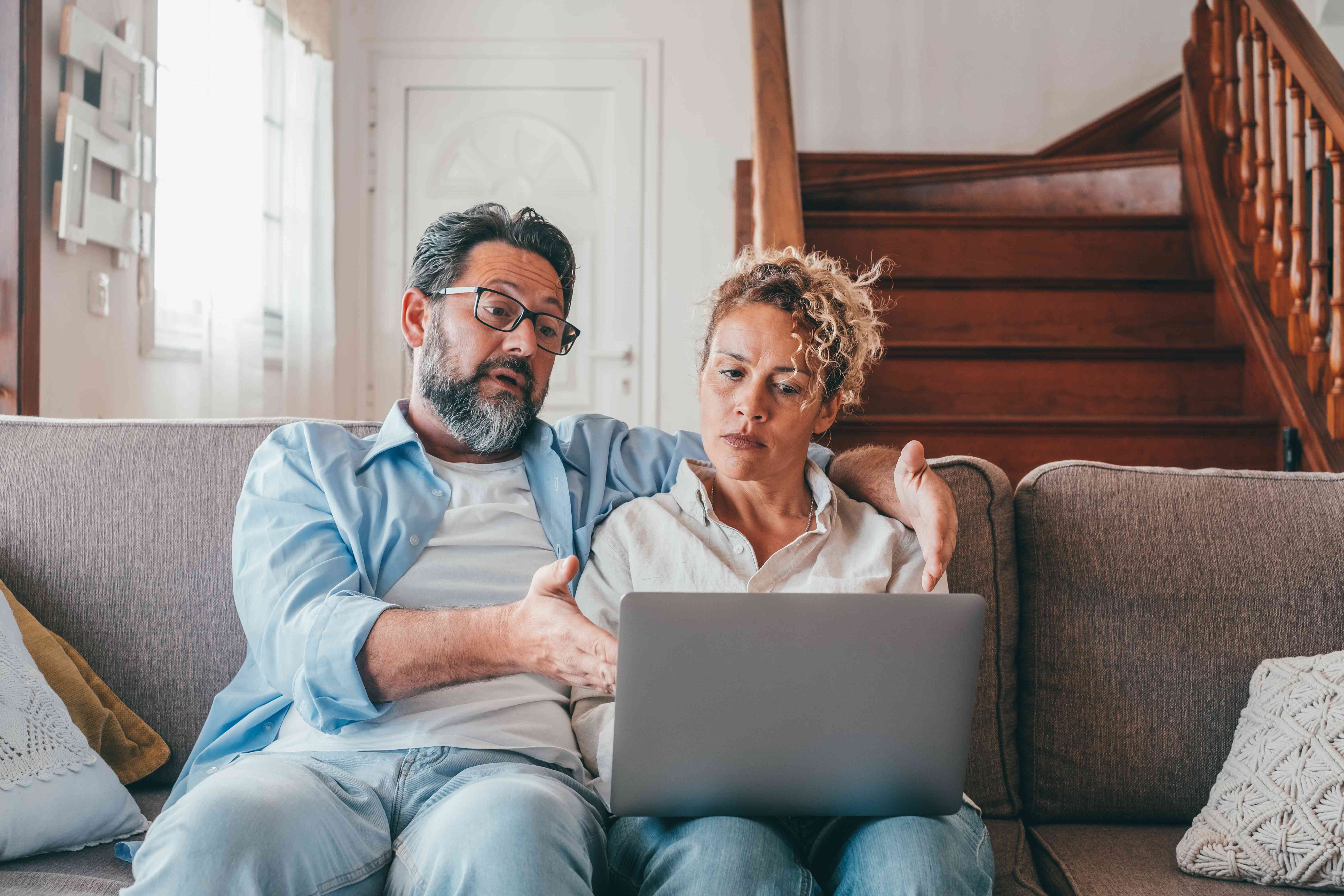 Couple in their 40s sitting together on their living room couch and looking at something on a laptop screen