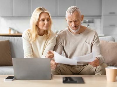 Older couple sitting on their living room couch and looking seriously at documents and a laptop.