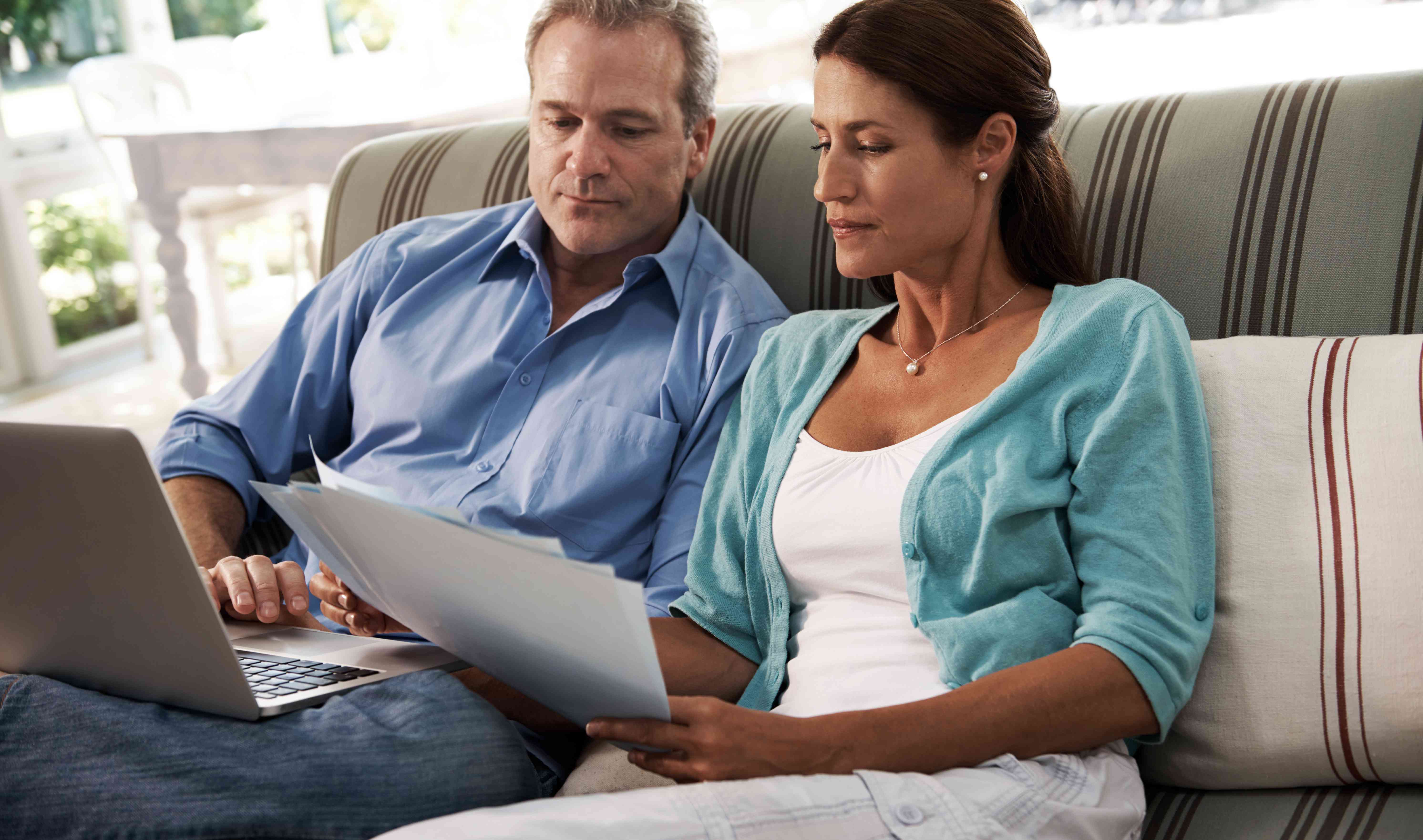 Couple in their 40s to 50s sitting on their living room couch and looking together at a laptop and documents