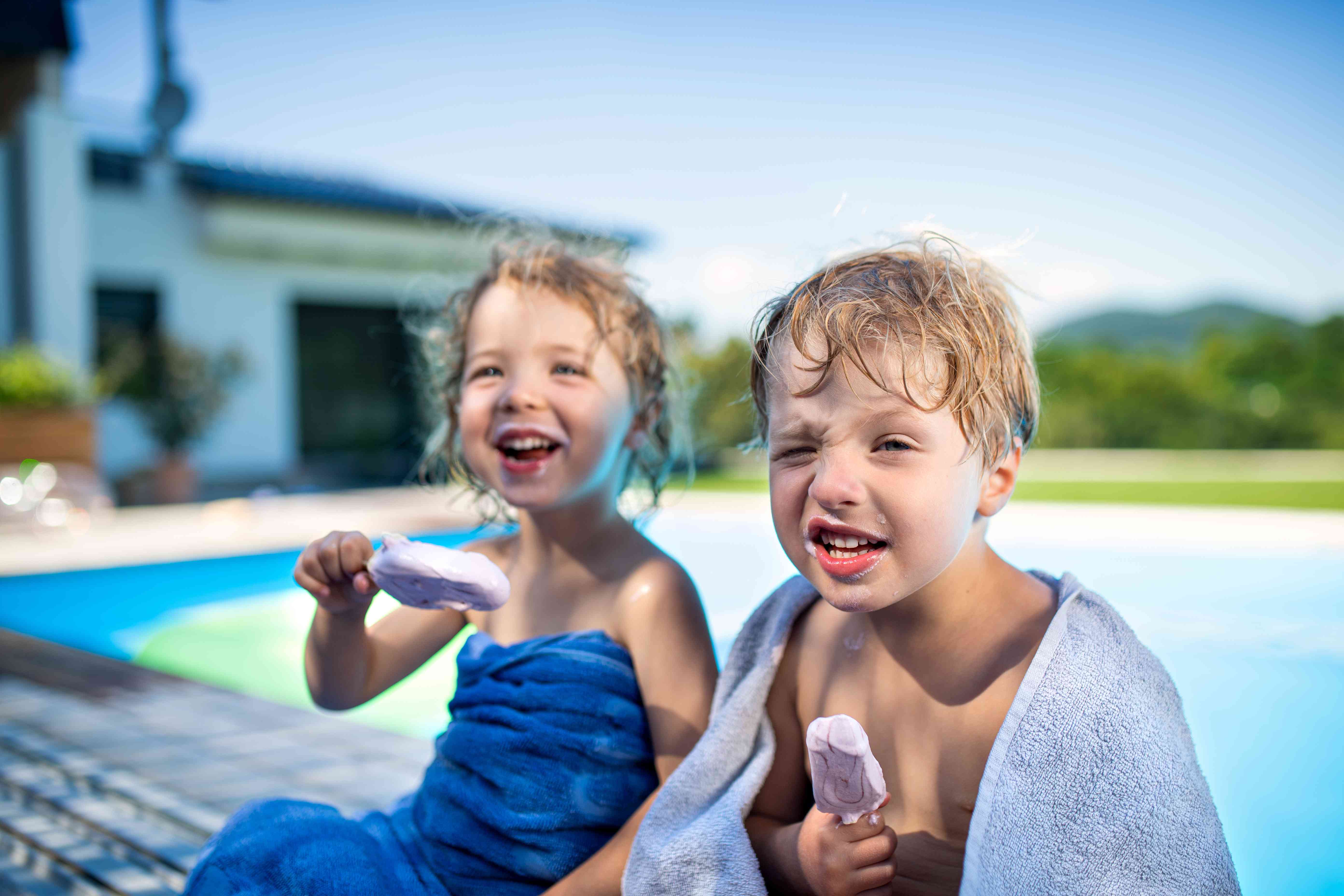 Two smiling young children sit poolside and enjoy ice cream pops.