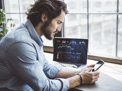 Young man sitting at a counter with a laptop showing market data and holding a smartphone