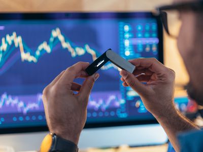 A man holds a cryptocurrency wallet while sitting in front of a computer screen displaying financial charts.