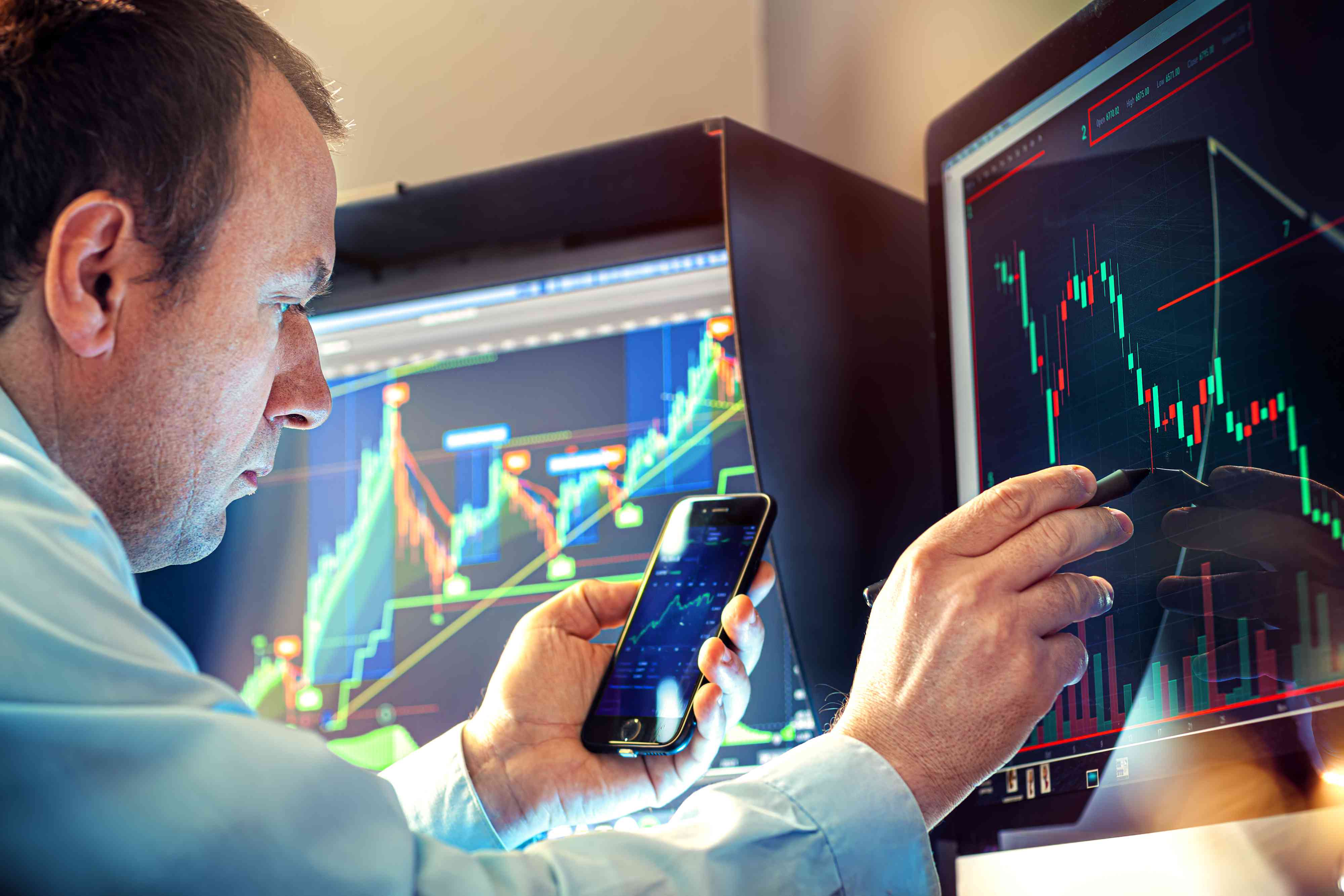 Stock trader with a smartphone in hand examines technical indicators in front of a computer monitor.