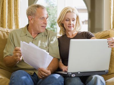 Older couple on living room couch, looking together at a laptop and financial documents