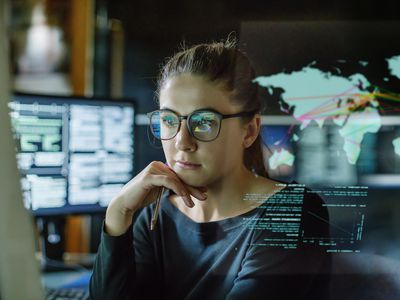 A woman wearing glasses sits at a computer, appearing behind a reflection displaying a world map.