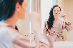 A woman stands in her bathroom brushing her teeth and looking at herself in the mirror while wearing a pink robe.