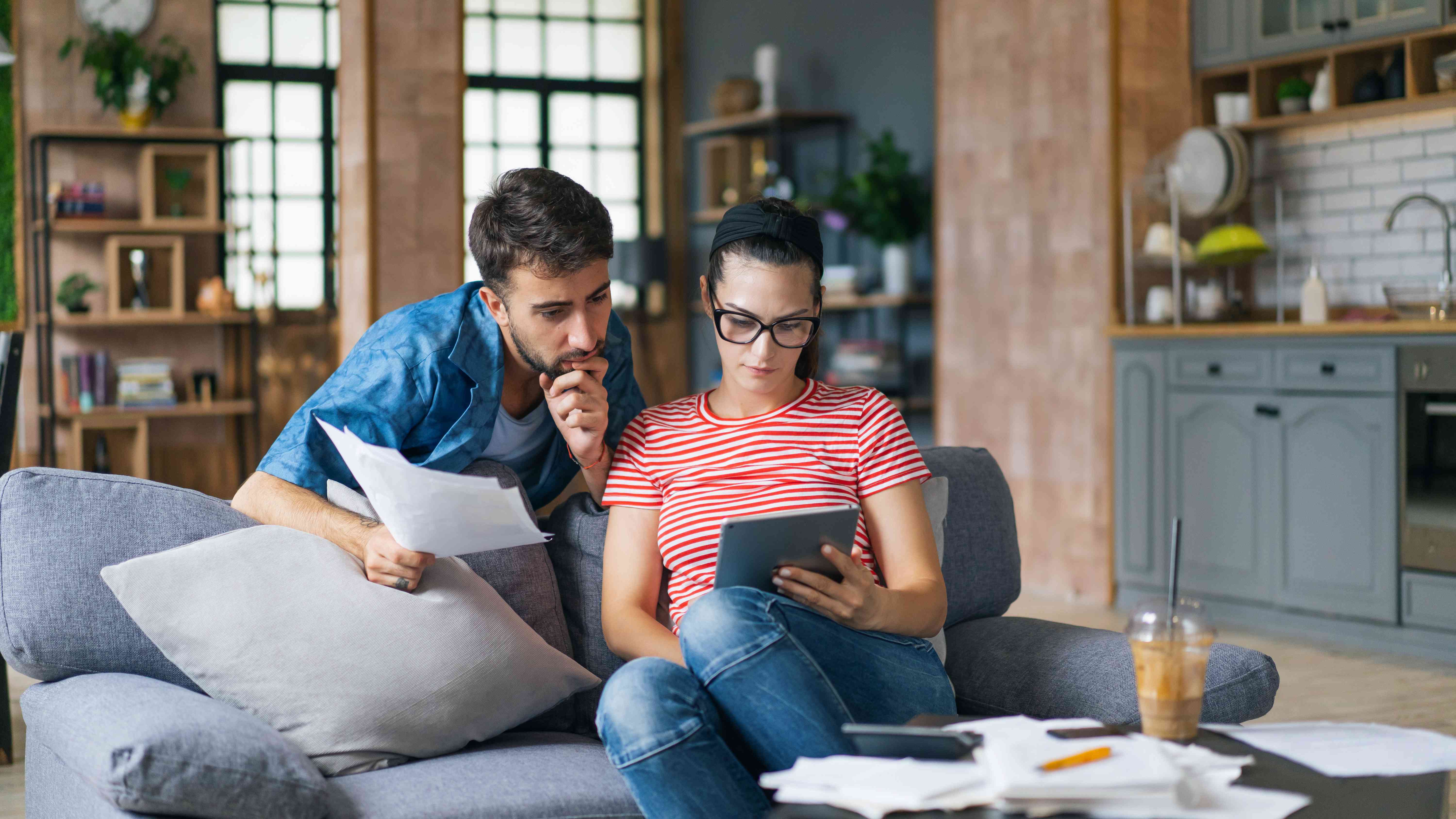 A man and a woman sit on a couch looking at documents and a tablet