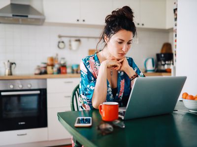 A woman in a blue and pink flowered robe sits at a kitchen table looking at a laptop. There is a phone, a red mug, and a bowl or oranges next to her.