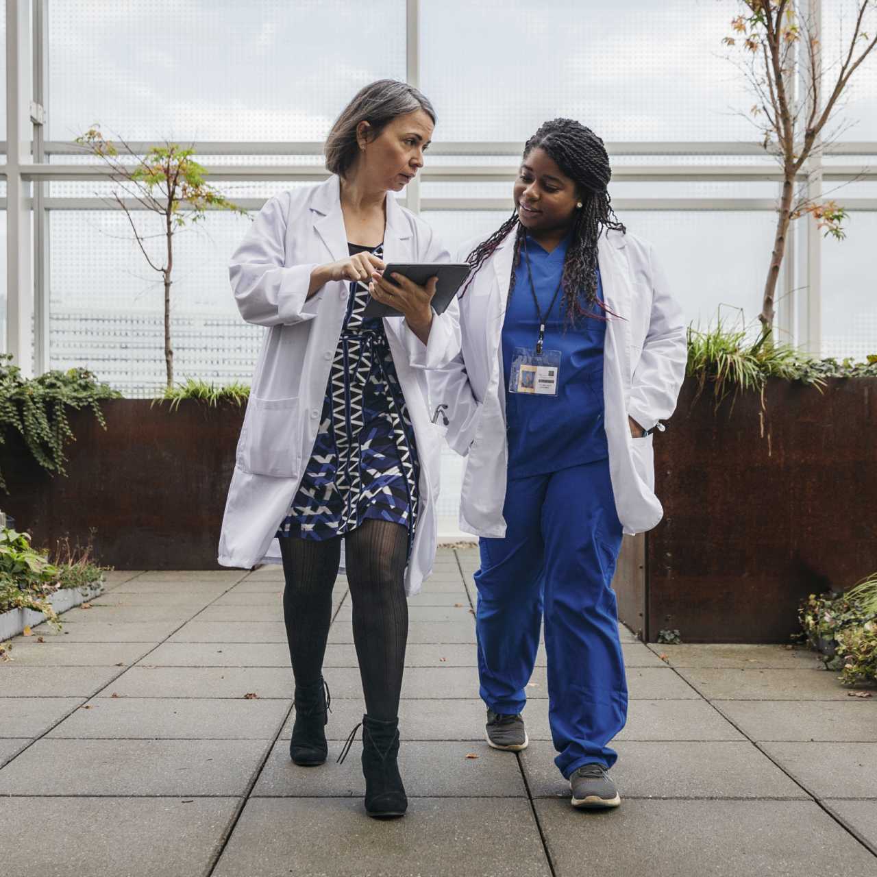 Two female healthcare workers in scrubs and white coats walking outside and talking.