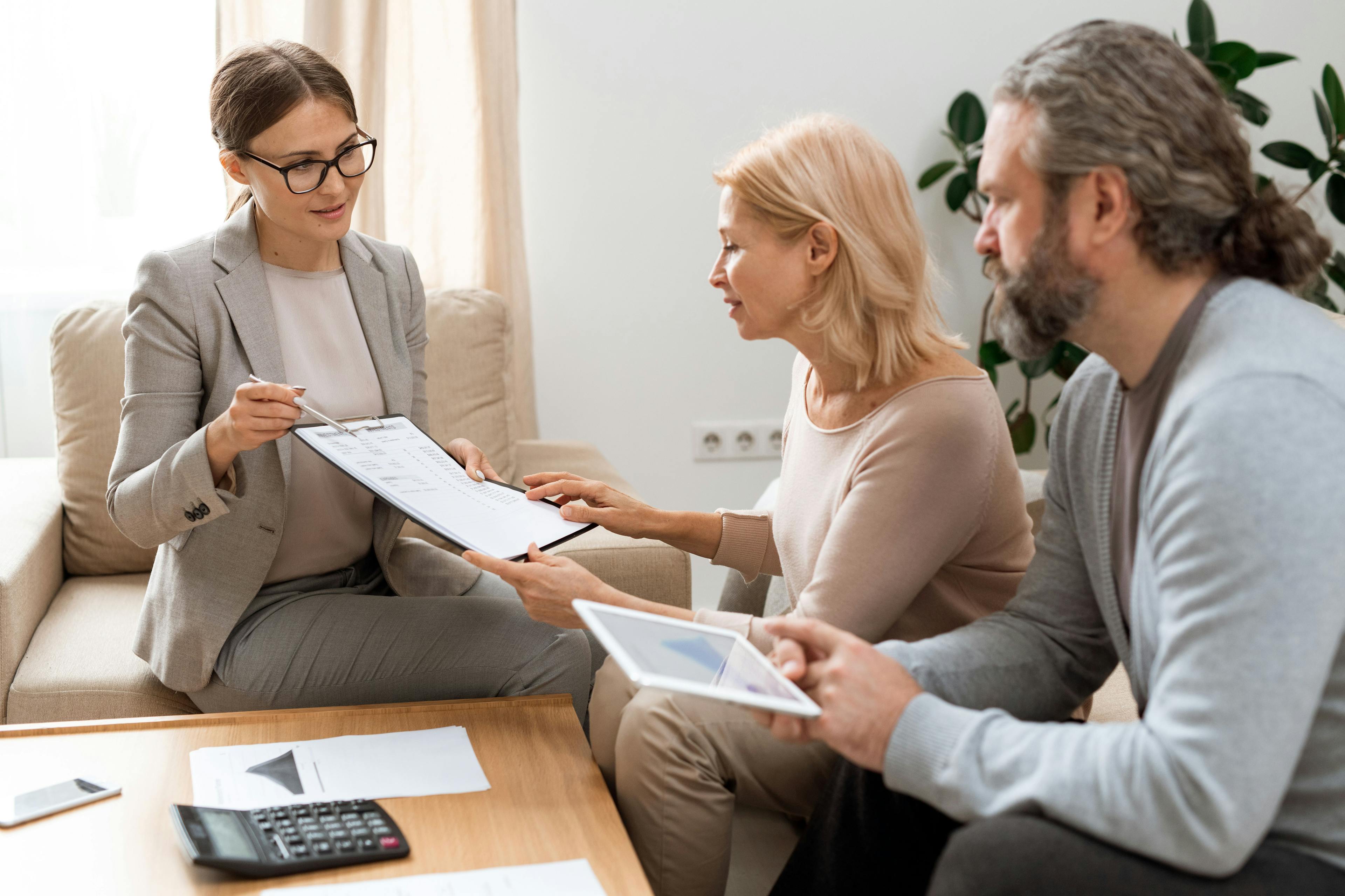 A young real estate agent from a search engine marketing company pointing at a document while explaining details to a middle-aged couple during a meeting in a cozy office setting.