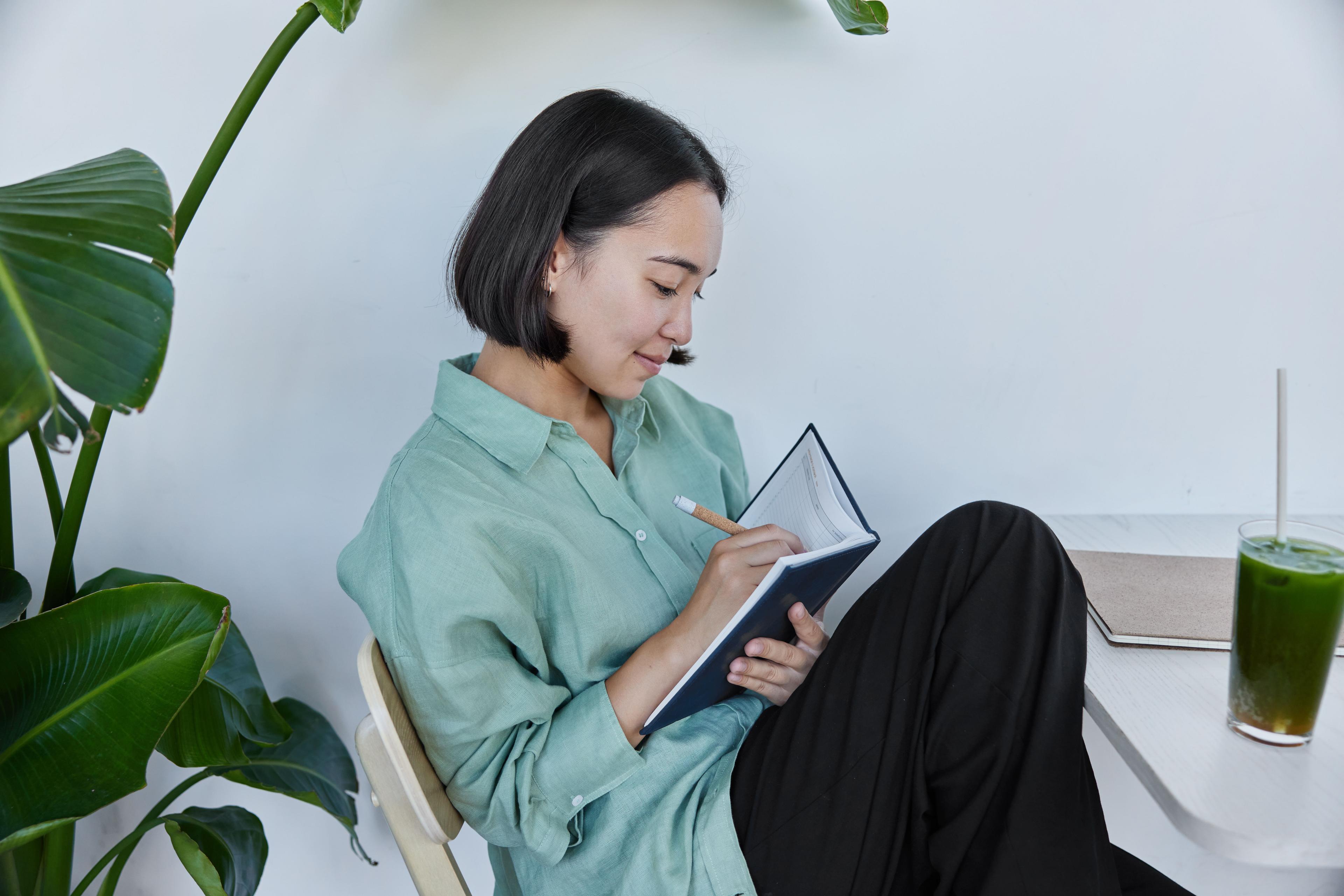 A woman sitting at a table with greenery around her, writing in a notebook. She appears focused and relaxed, which could symbolize the planning and strategy phase of search engine optimization marketing.