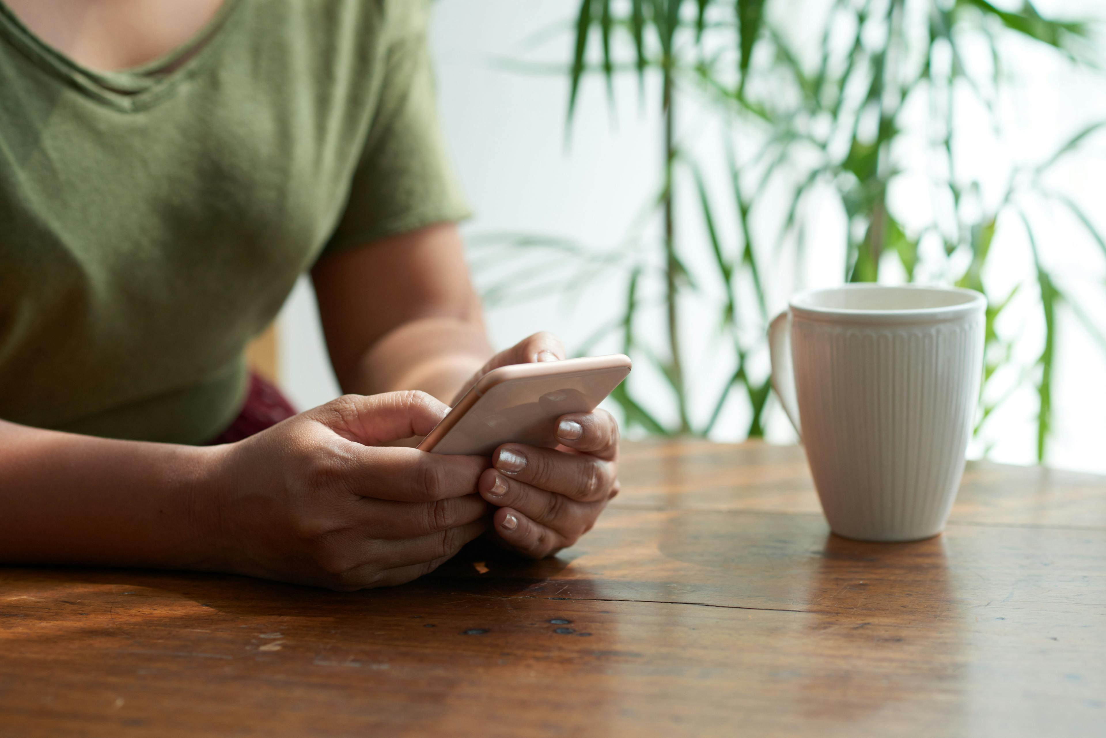 A person sitting at a table, holding a smartphone and a coffee cup nearby, with a plant in the background. This scene represents "small business marketing consultants" staying connected with clients and partners through digital communication and mobile devices.