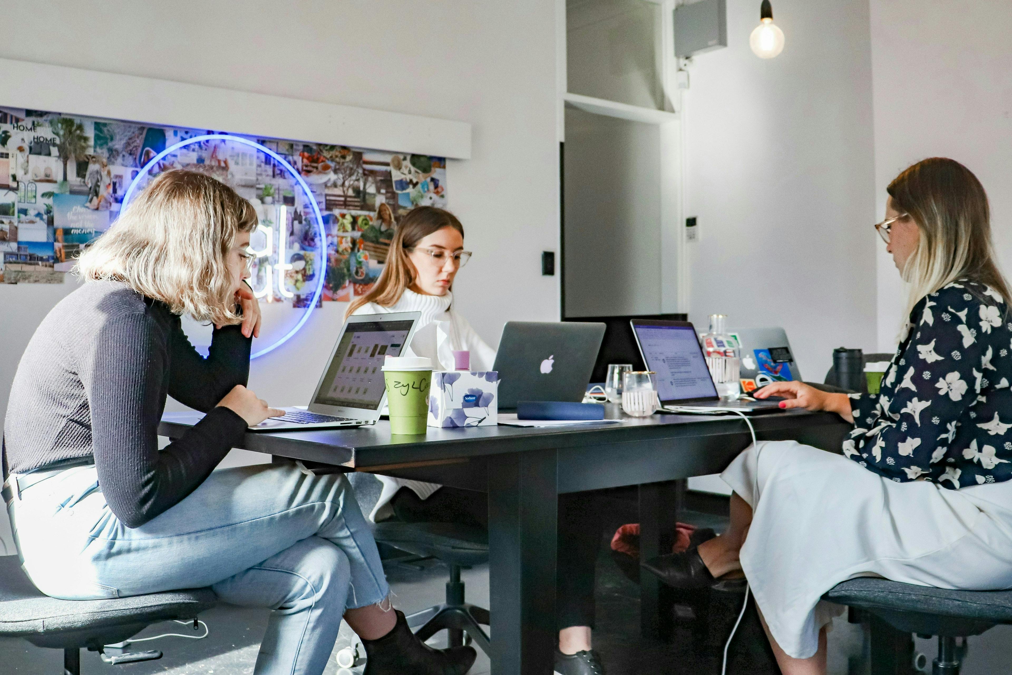 Three women are working on laptops around a conference table in a modern office setting. The workspace features a neon sign on the wall and various decorative elements, creating a creative and collaborative atmosphere. This image is ideal for illustrating the use of AI image generators like Bing in a professional environment.