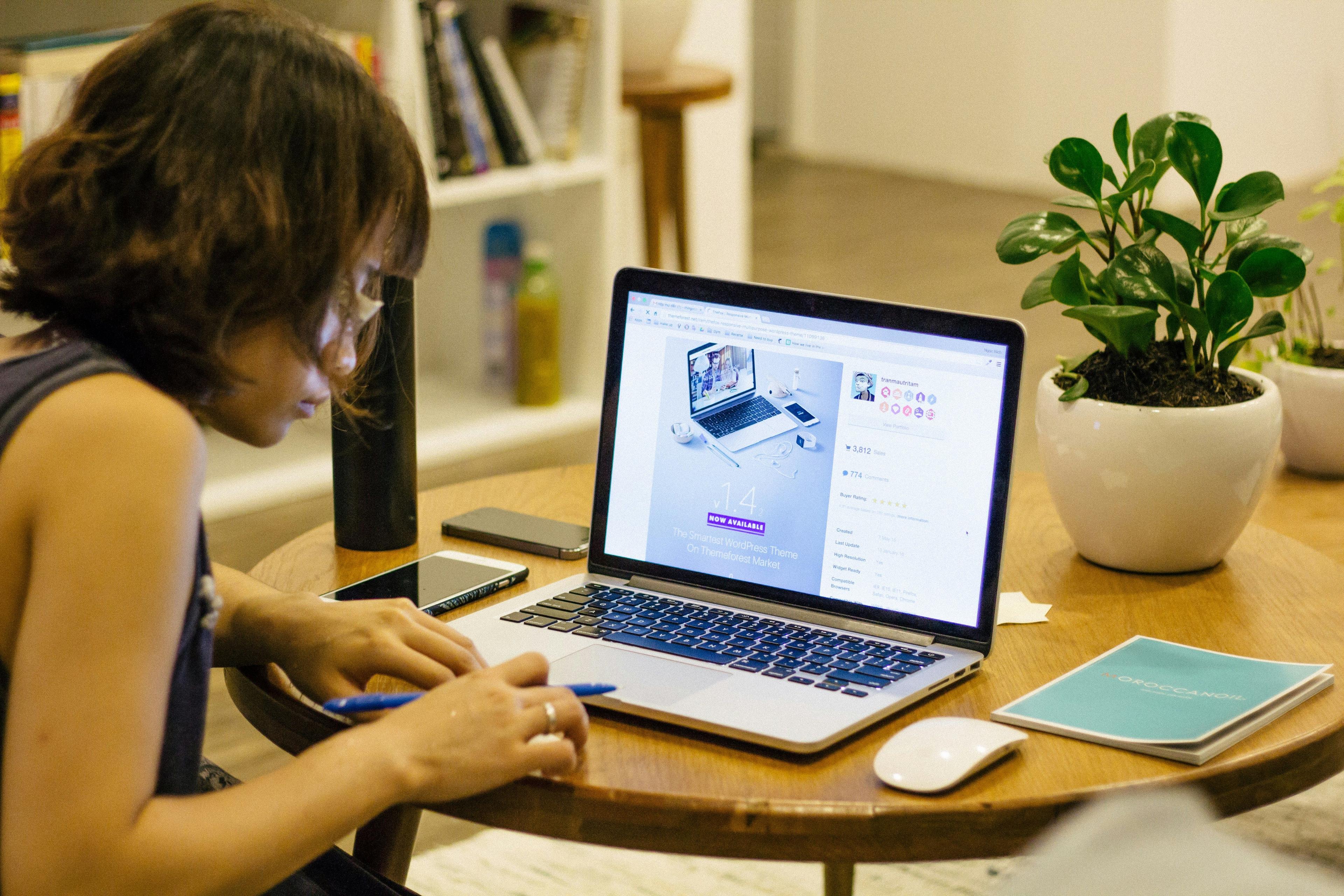 A woman working intently on her laptop at a wooden table, with the screen displaying a webpage related to WordPress themes. The workspace includes a phone, a notebook, a pen, and a potted plant, creating a cozy and productive environment. This image is perfect for illustrating the use of AI copywriting tools like WriterZen.