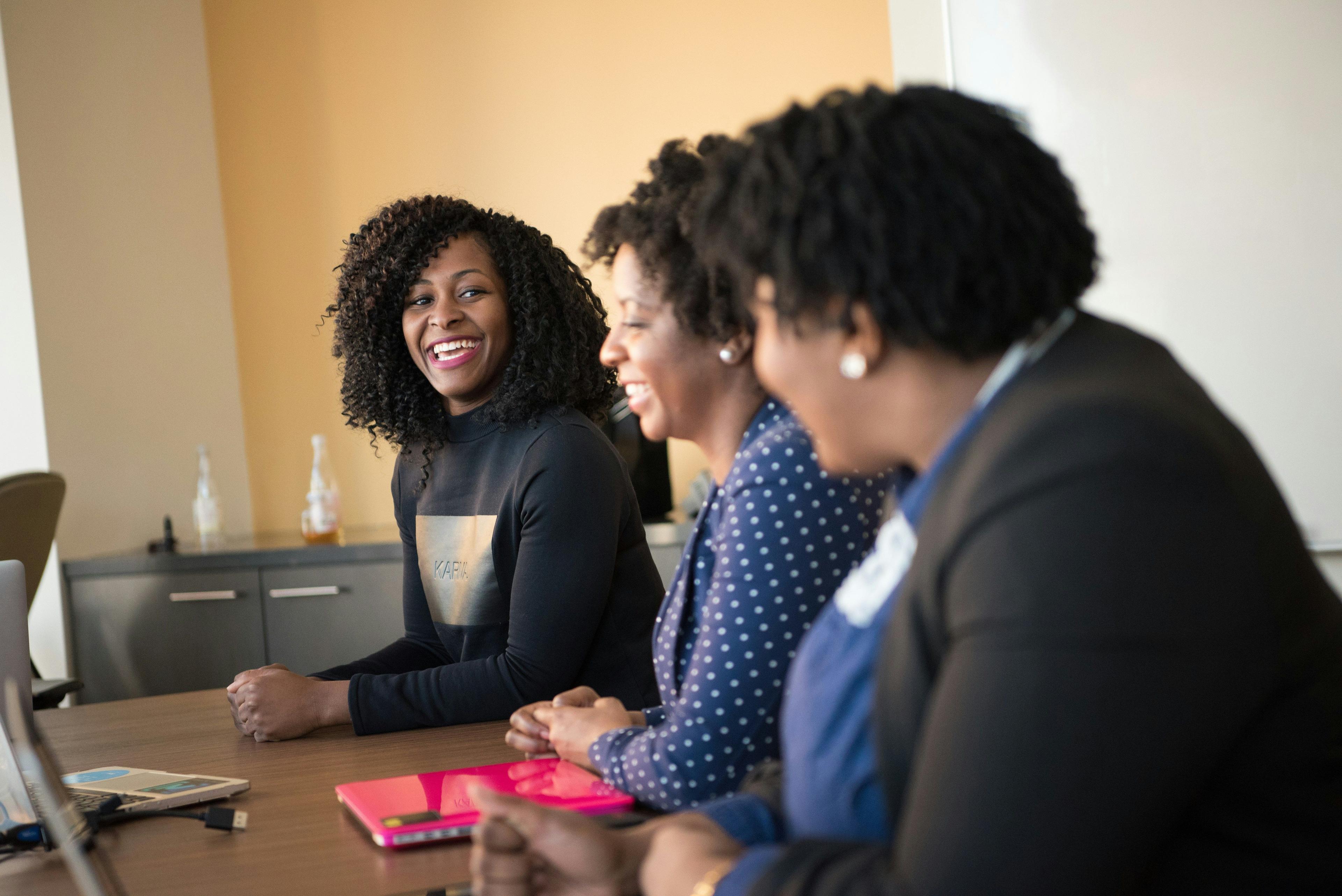 Three women of diverse backgrounds sitting at a conference table, engaging in a lively discussion, representing a team brainstorming session for social media marketing strategies.