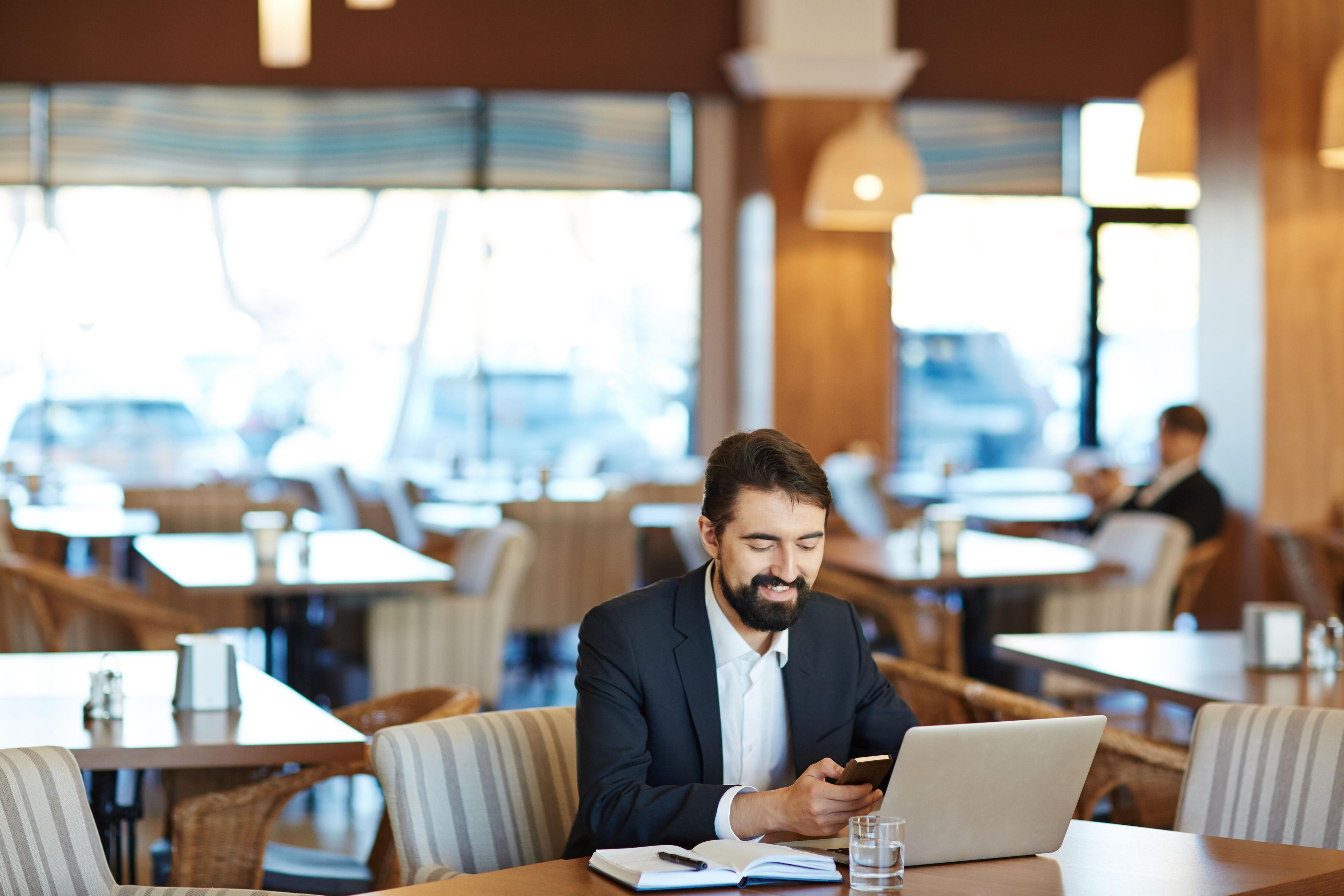 Cheerful businessman working in a cafe, looking at his phone and laptop while smiling, illustrating a scenario related to social media ad cost.